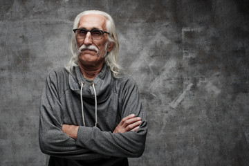 Old Caucasian gray-haired cool grandfather with glasses incredulously looks into the camera with crossed arms and serious face with copy space in studio