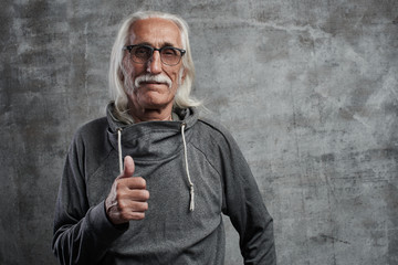 Modern gray-haired caucasian old man with a mustache shows thumb up and smiles, joyful happy face against gray wall in studio