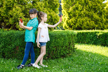 Two cute girls of school age photograph themselves on the phone near the green trees. Children are having fun