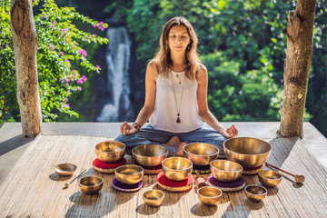 Woman playing on Tibetan singing bowl while sitting on yoga mat against a waterfall. Vintage tonned. Beautiful girl with mala beads meditating