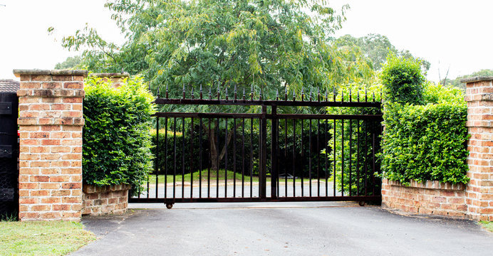 Black metal driveway property entrance gates set in brick fence with garden shrubs and trees in background