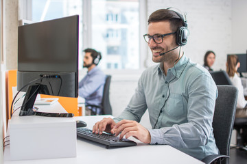 Smiling handsome male customer support phone operator with headset working in call centre. Group of...