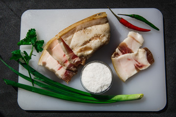 Sliced pieces of lard with layers of meat on a white Board decorated with herbs and salt in a salt shaker. Appetizing still life from a fresh piece of fat on the table