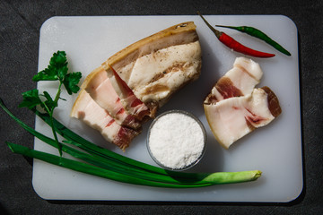 Sliced pieces of lard with layers of meat on a white Board decorated with herbs and salt in a salt shaker. Appetizing still life from a fresh piece of fat on the table