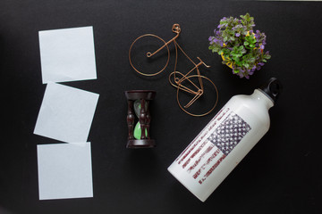 water bottle, hourglass and flower with toy bike on black background. Flat Lay with sand, dust and...
