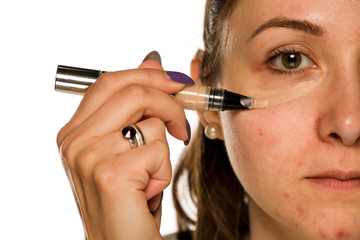 Young woman applying concealer on her face on white background