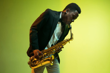 Young african-american jazz musician playing the saxophone on gradient yellow-green studio...