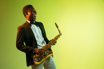 Fototapeta na wymiar Young african-american jazz musician playing the saxophone on gradient yellow-green studio background. Concept of music, hobby, festival. Joyful attractive guy improvising. Colorful portrait of artist