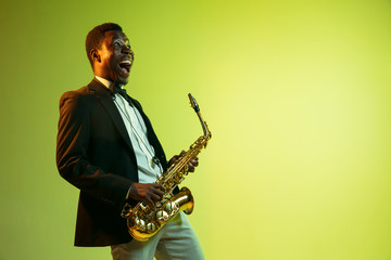 Naklejka premium Young african-american jazz musician playing the saxophone on gradient yellow-green studio background. Concept of music, hobby, festival. Joyful attractive guy improvising. Colorful portrait of artist