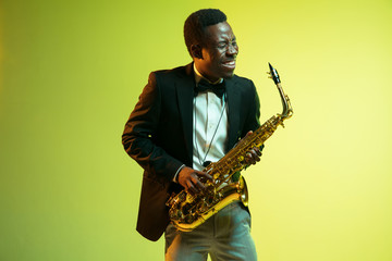 Naklejka premium Young african-american jazz musician playing the saxophone on gradient yellow-green studio background. Concept of music, hobby, festival. Joyful attractive guy improvising. Colorful portrait of artist