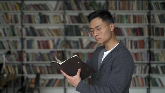 Middle shot of concentrated serious black hair asian young man in grey jacket and big eyeglasses standing nodding making notes in notepad. Bookshelves