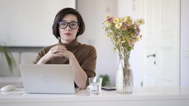 Gimbal shot of serious asian business woman with short haircut sitting looking at the camera hands crossed. Glass of water on table and a vase with flowers in white interior home office