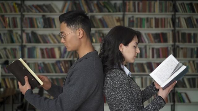 Multi ethnic couple asian man and oriental woman profile holding flipping through books standing back to back reading in library on background of bookshelves