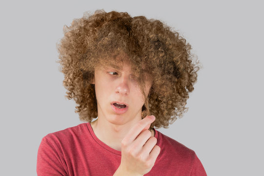 Portrait Of A Young Curly European Man Frightened Looks At His Long Hair. Holds A Curl Of Hair With His Fingers. Very Lush Male Guy Hair. Curling Hair For Men. Isolated On Gray Background