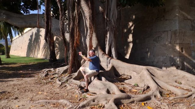 A male tourist sits on a big tree root in front of the wall of the University of the Spanish port city Cartagena and takes a picture with his smartphone. The man is enjoying the sunshine.