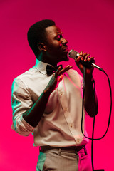 Young african-american jazz musician singing a song on gradient pink-red background in neon light. Concept of music, hobby, festival, open-air. Joyful guy improvising. Colorful retro portrait of