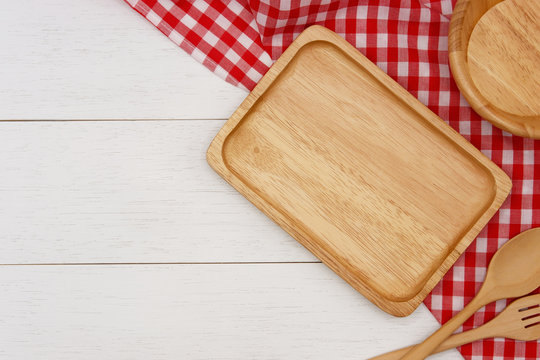 Empty rectangle wooden plate with spoon, fork and red gingham tablecloth on white wooden table. Top view image with copy space.