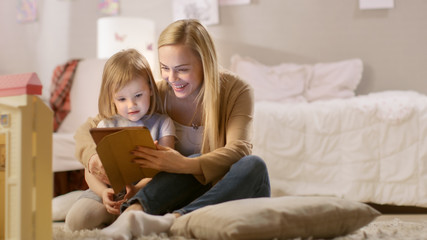 Beautiful Mother and Her Little Daughter Have Good Time Reading Children's Books on a Tablet Computer. Sunny Living Room.