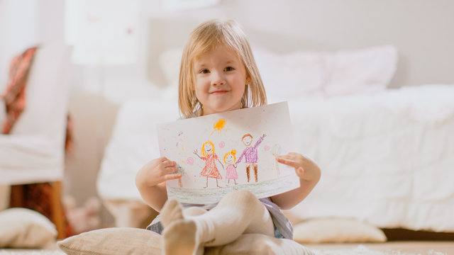 Cute Young Smiling Girl Sitting on Pillows and Shows Drawing of Her Family. Sunny Living Room.