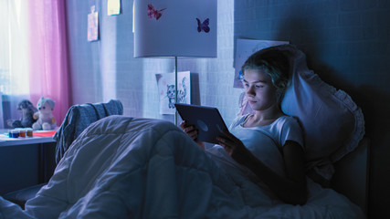 Fototapeta premium Young Teenage Girl Lies in Her Bed at Night With Tablet Computer. Watches TV Show and Smiles.
