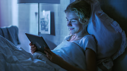 Young Teenage Girl Lies in Her Bed at Night With Tablet Computer. Watches TV Show and Smiles.