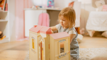 In the Childrens Room: Cute Happy Little Girl Playing with Doll House and Sitting on a Carpet. Sunny Living Room with Children Having Fun. 