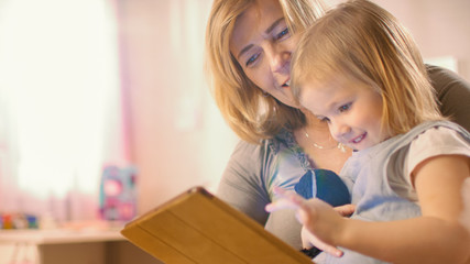 Beautiful Mother and Her Cute Little Daughter Sitting on the Floor and Reading Children's Books on a Tablet Computer. Sunny Living Room. 