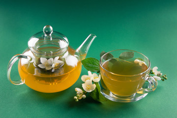 Glass tea cup and pot filled with jasmine tea and jasmine stem on green background. Side view. Soft focus