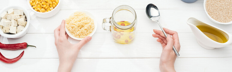 cropped view of woman holding bowl with couscous near glass jar on wooden white table, panoramic shot