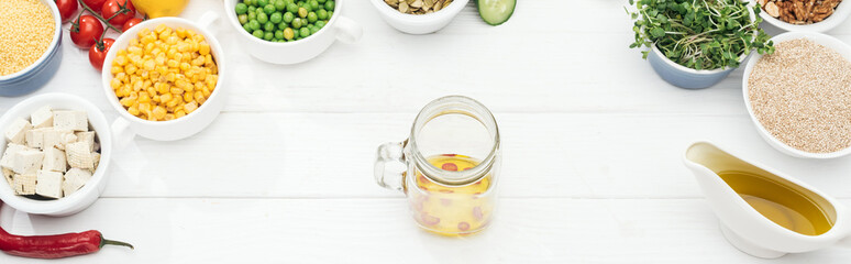 top view of glass jar with oil and chili pepper near fruits and vegetables near glass jars on wooden white table, panoramic shot