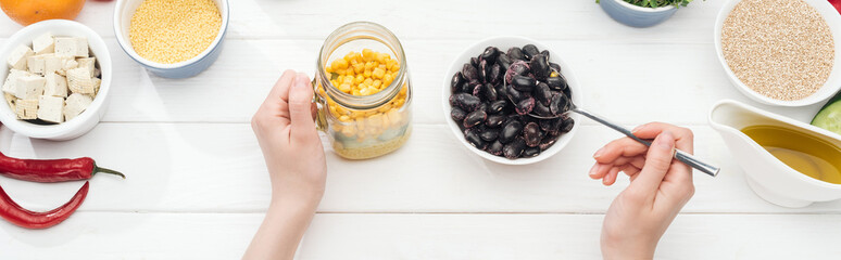 cropped view of woman adding beans in jar with salad on wooden white table, panoramic shot