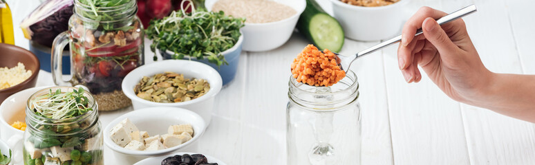 cropped view of woman adding couscous in glass jar on wooden white table, panoramic shot