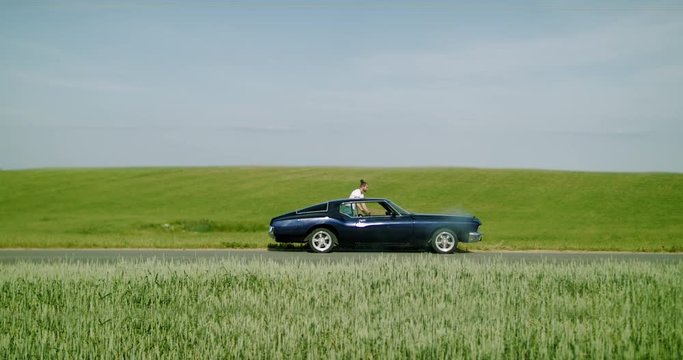 Caucasian male standing near overheated old vintage car in the field, bright sunlight, smoke from under the hood. 4K UHD RAW graded footage