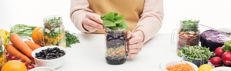 partial view of woman in apron holding glass jar with salad and green leaves on wooden table isolated on white, panoramic shot