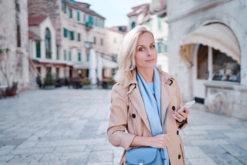 Fototapeta premium Travel and technology. Happy young woman in coat using smartphone walking by Split Old Town.