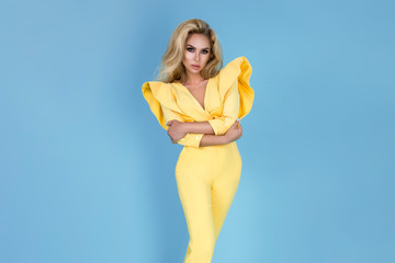 Elegant blonde woman in stylish yellow jumpsuit and fashionable accessories on color background. Beautiful fashion model on blue background.