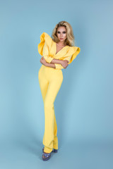 Elegant blonde woman in stylish yellow jumpsuit and fashionable accessories on color background. Beautiful fashion model on blue background.