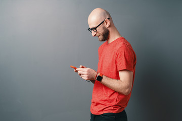 Naklejka premium Handsome hairless Caucasian man with beard, glasses, red T-shirt talking on the phone on gray studio background