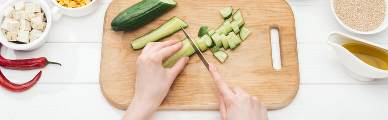 cropped view of woman chopping cucumber on wooden white table, panoramic shot