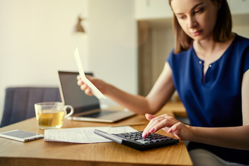 Thoughtful young woman using a laptop computer sitting at her kitchen holding utility bill and bank...