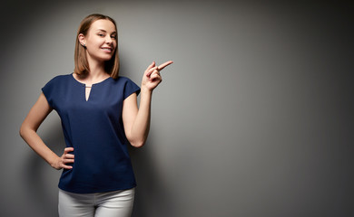 Copy space for avertisement. Portrait of happy smiling young woman pointing away against grey wall.