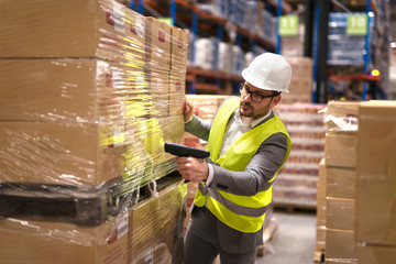 Working at warehouse. Male warehouse worker using bar code scanner to analyze newly arrived goods for further placement in storage department. Employee organizing goods distribution to the market. - obrazy, fototapety, plakaty