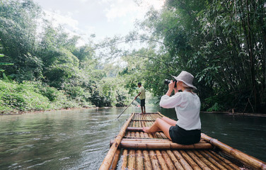Traveling by Thailand. Pretty young woman taking photo sailing jungle river on traditional bamboo raft.