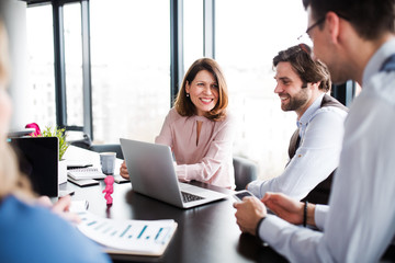 A group of young business people with laptop sitting in an office, talking.