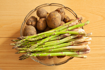 Fresh new potatoes with peel on and green asparagus in a glass bowl. Top view close up image on wooden background.