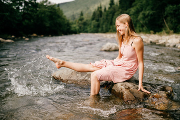 Young happy girl kicking foot in river and splashing water.