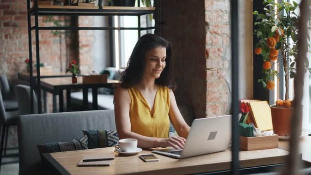 Attractive girl is using laptop in cafe typing smiling working in cafe alone enjoying freelance work. Modern technology, happy youth and devices concept.