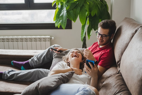 A young couple rests on the couch at home while looking at the mobile phone