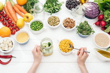 cropped view of woman adding corn to jar with cucumbers on wooden white table