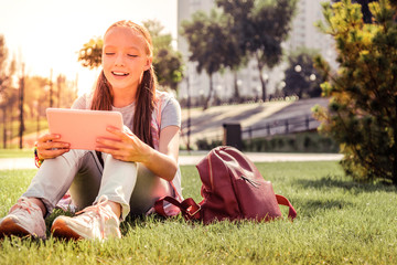 Smiling young lady chilling on a green grass and checking tablet